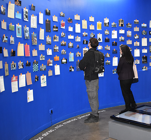 Dos personas observan la pared azul de la entrada a la exposición cuando escribo obras, mismis manos piensan libros